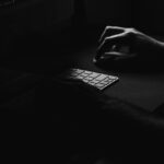Moody black and white photo of hands typing on a keyboard in the dark, creating a mysterious atmosphere.